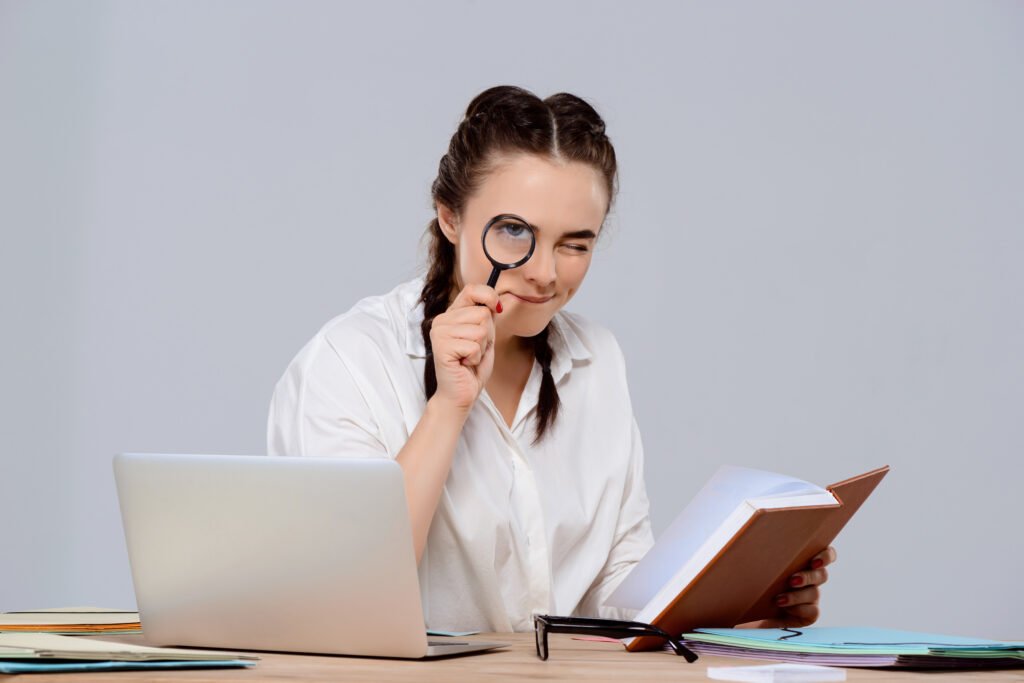 Young beautiful businesswoman sitting at workplace, holding book and magnifier.