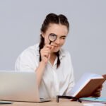 Young beautiful businesswoman sitting at workplace, holding book and magnifier.